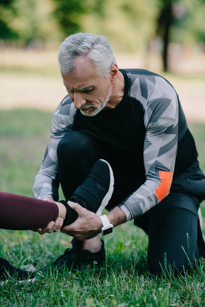 Schöner älterer Mann berührt verletztes Bein einer Sportlerin, die auf Rasen im Park sitzt - Foto, Bild