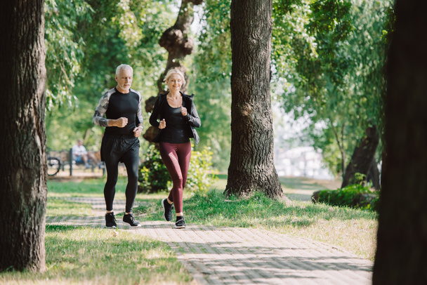 selective focus of smiling mature sportsman and sportswoman running together in park - Photo, Image