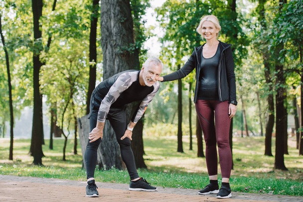 tired mature sportsman and smiling sportswoman looking together while standing in park - Foto, Imagem
