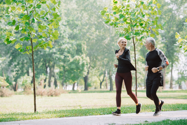 χαρούμενο ώριμο αθλητή και αθλητική γυναίκα τρέξιμο μαζί στο ηλιόλουστο πάρκο - Φωτογραφία, εικόνα