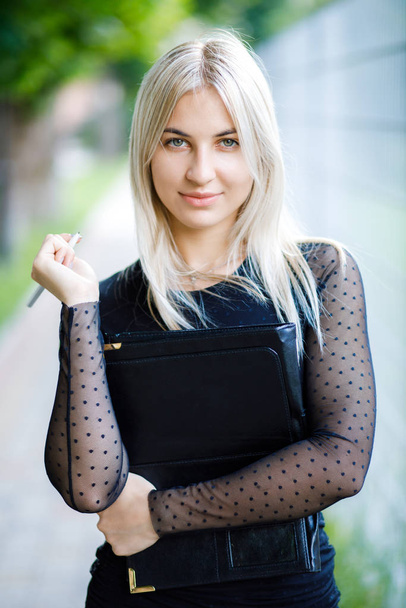 A young beautiful blonde girl wearing a classic black dress holds a folder of papers in her hands. Business clothing style. - Foto, imagen