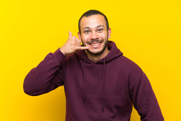 Κολομβιανός άνθρωπος με φούτερ πάνω από κίτρινο τοίχο κάνοντας χειρονομία τηλέφωνο. Καλέστε με πίσω πινακίδα - Φωτογραφία, εικόνα