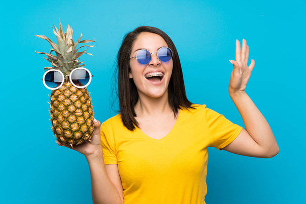 Young Mexican woman over isolated blue background holding a pineapple with sunglasses - Photo, Image