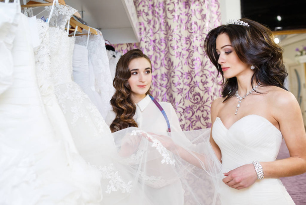 Female trying on wedding dress in a shop with women assistant. - Photo, Image