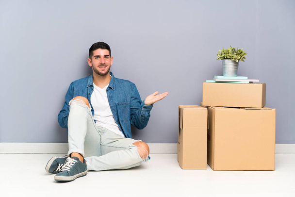 Handsome young man moving in new home among boxes holding copyspace imaginary on the palm - Photo, Image