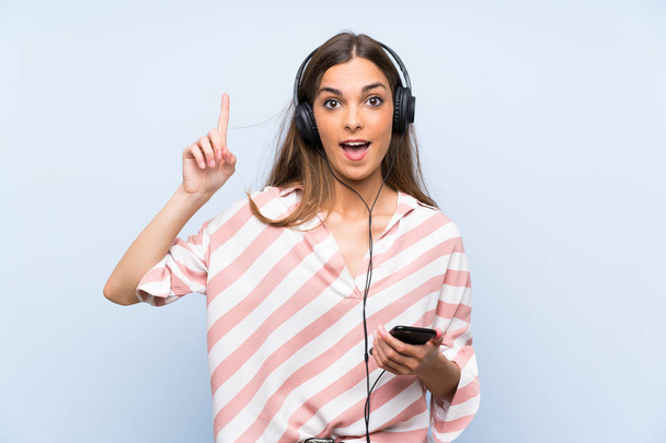 Nuori nainen kuuntelee musiikkia mobiili yli eristetty sininen seinä aikoo toteuttaa ratkaisun nostaessaan sormea ylös
 - Valokuva, kuva