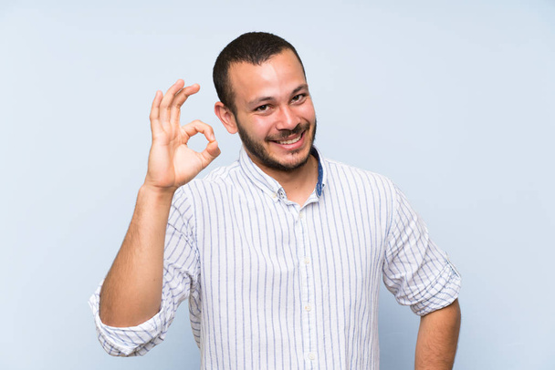 Kolumbijski człowiek na białym tle niebieską ścianę pokazując OK znak palcami - Zdjęcie, obraz