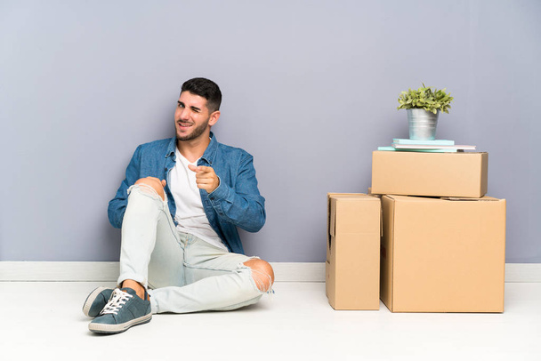 Handsome young man moving in new home among boxes points finger at you - Foto, afbeelding