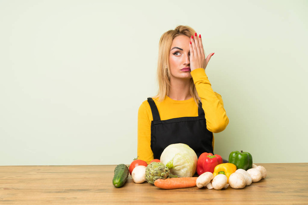 Young blonde woman with lots of vegetables covering a eye by hand - Photo, Image
