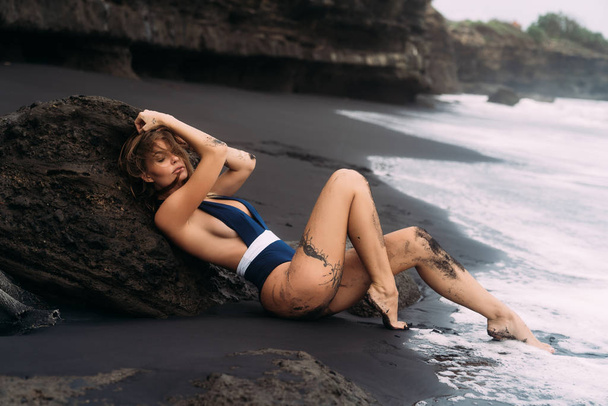 Sexy girl with big breasts in blue swimwear relaxing at beach with black sand. - Zdjęcie, obraz