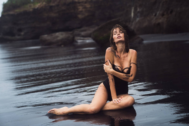 Sensual sexy girl in swimsuit with wet hair sits on black sand beach - Фото, зображення
