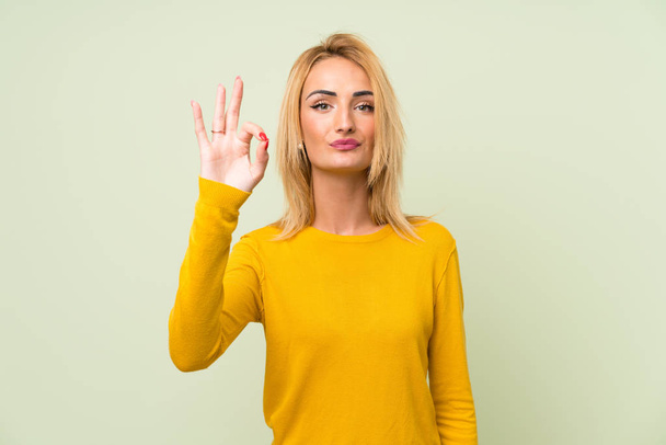 Jonge blonde vrouw over geïsoleerde groene achtergrond toont een OK teken met vingers - Foto, afbeelding