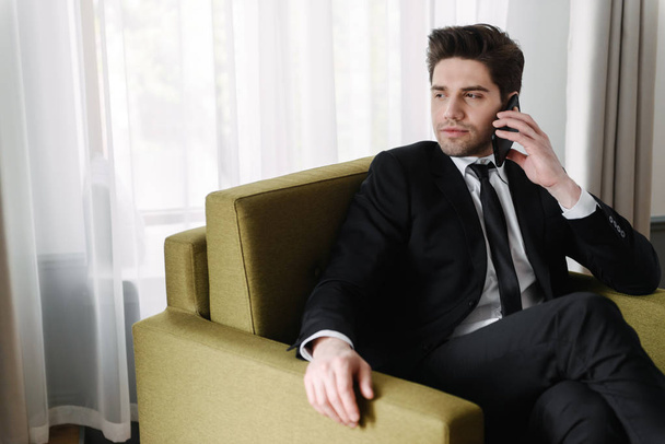 Photo of brooding handsome businessman talking on cellphone while sitting on armchair in hotel apartment - Photo, Image