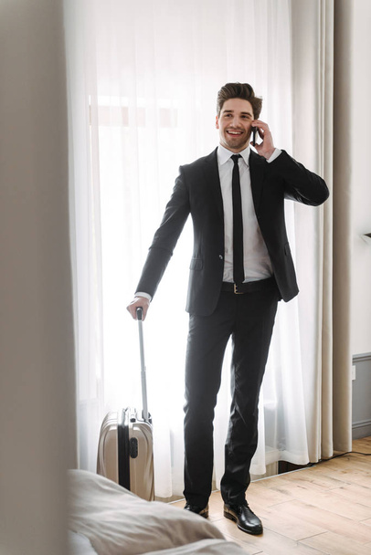Photo of smiling young businessman talking on cellphone and holding luggage in hotel apartment - Фото, зображення