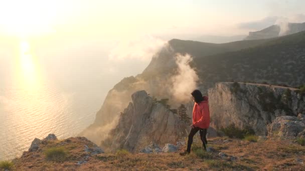 若い男はクリミアの高い山から海の上に美しい劇的なカラフルな夕日を観察崖の端に沿って歩きます. - 映像、動画