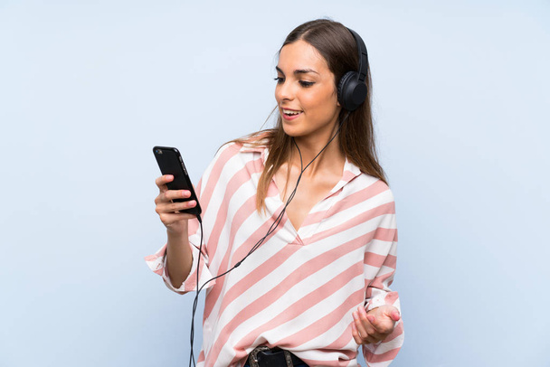 Mladá žena poslouchá hudbu s mobilem přes izolovanou modrou zeď - Fotografie, Obrázek