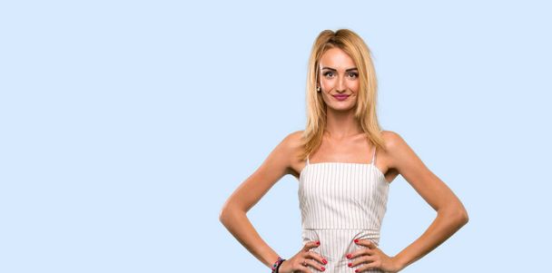 Young blonde woman posing with arms at hip and smiling over isolated blue background - Photo, Image