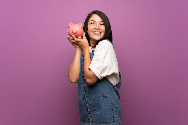 Young Mexican woman over isolated background holding a big piggybank - 写真・画像
