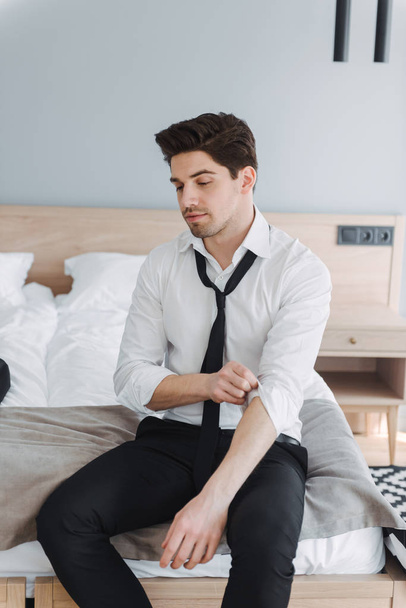 Photo of caucasian young businessman looking downward while sitting on bed in hotel apartment - 写真・画像