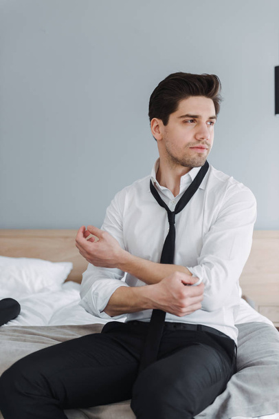 Photo of young brunette businessman looking aside while sitting on bed in hotel apartment - Photo, Image