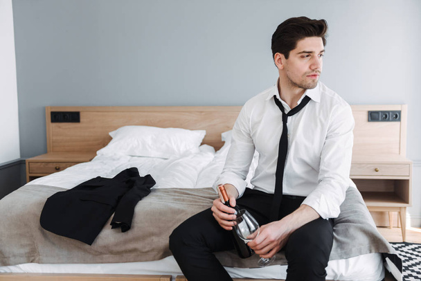 Foto von brünetten schönen Geschäftsmann hält Flasche Wein und Glas, während er auf dem Bett in der Hotelwohnung sitzt - Foto, Bild