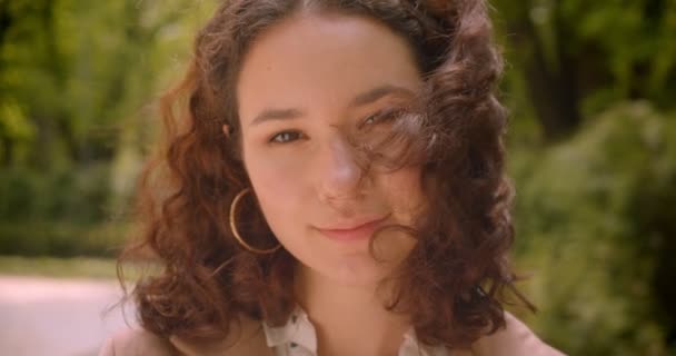 Closeup portrait of young pretty long haired curly caucasian female cheerfully looking at camera standing outdoors in the garden - Materiał filmowy, wideo