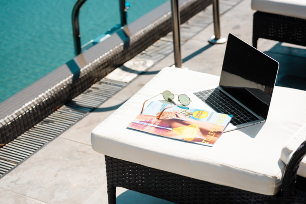 portátil con pantalla en blanco, revista y gafas de sol en la cama de sol cerca de la piscina
 - Foto, Imagen