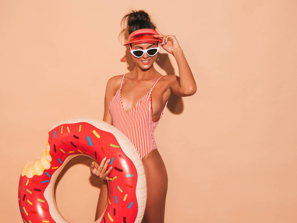 Young beautiful sexy smiling hipster woman in sunglasses.Girl in summer swimwear bathing suit with donut lilo inflatable mattress.Positive female going crazy.Near beige wall in transparent visor cap - Foto, Bild