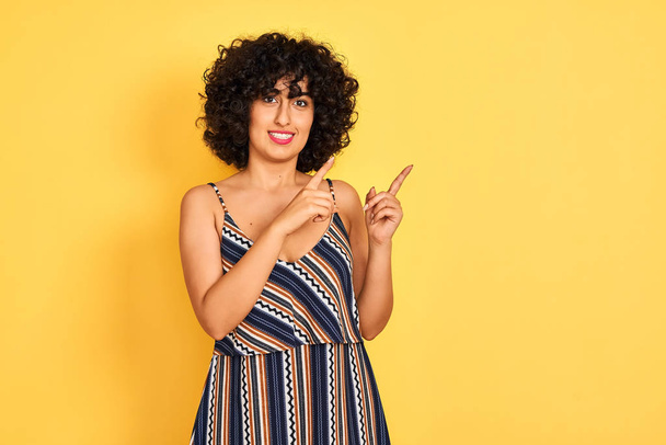 Femme arabe aux cheveux bouclés portant une robe rayée colorée sur fond jaune isolé souriant et regardant la caméra pointant avec deux mains et doigts sur le côté
. - Photo, image