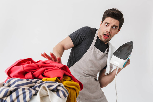 Confused houseman househusband holding iron looking at clothes isolated over white wall background. - Foto, Imagen
