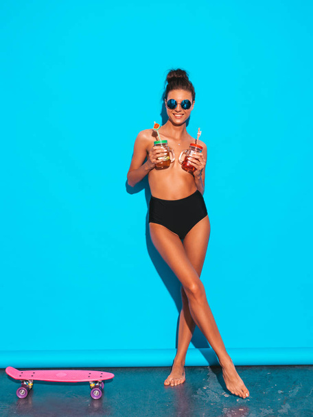 Young beautiful sexy smiling hipster woman in sunglasses.Girl in summer  swimwear bathing suit.Posing near blue wall,drinking fresh cocktail smoozy drink.Covers her chest by glass jar - Fotografie, Obrázek