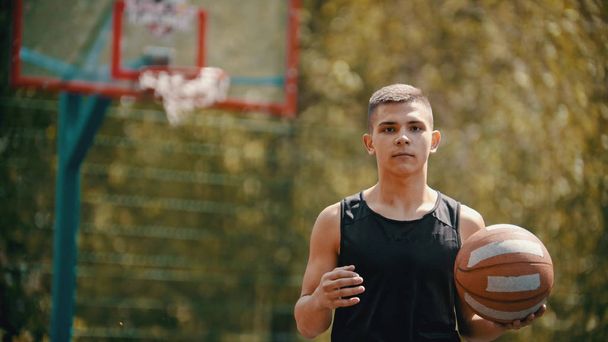 Ένας νέος αθλητικός άνθρωπος στέκεται σε ένα αθλητικό έδαφος και κρατώντας μια μπάλα του μπάσκετ - Φωτογραφία, εικόνα