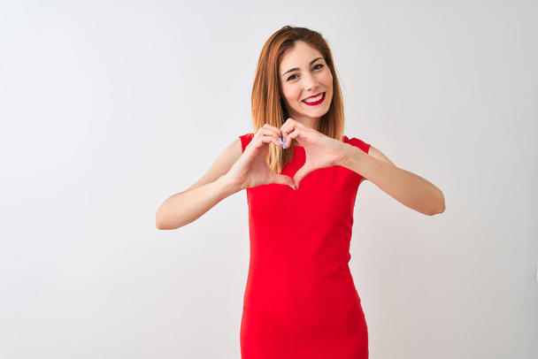 Κοκκινομάλλα επιχειρηματίας φορώντας κομψό κόκκινο φόρεμα στέκεται πάνω από απομονωμένο λευκό φόντο χαμογελώντας στην αγάπη κάνει το σχήμα σύμβολο της καρδιάς με τα χέρια. Ρομαντική ιδέα. - Φωτογραφία, εικόνα