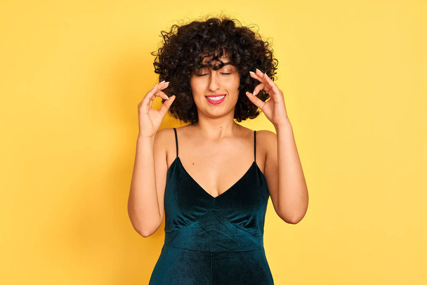 Jeune femme arabe aux cheveux bouclés portant une robe élégante sur fond jaune isolé se détendre et sourire les yeux fermés en faisant geste de méditation avec les doigts. Concept de yoga
. - Photo, image