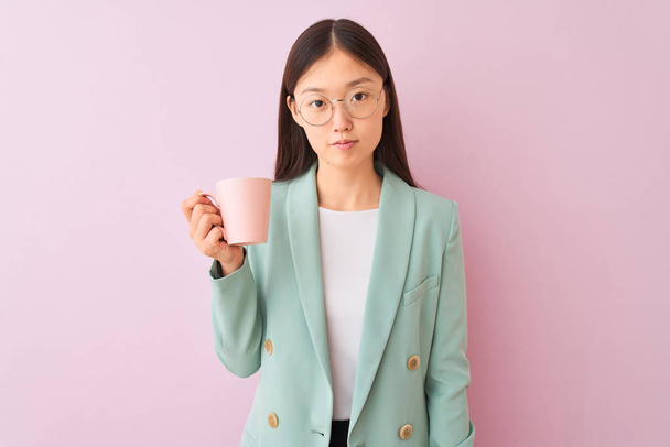 junge chinesische Geschäftsfrau trägt Gläser Kaffee trinken über isolierten weißen Hintergrund mit einem selbstbewussten Gesichtsausdruck auf Smart Gesichtsdenken ernst - Foto, Bild