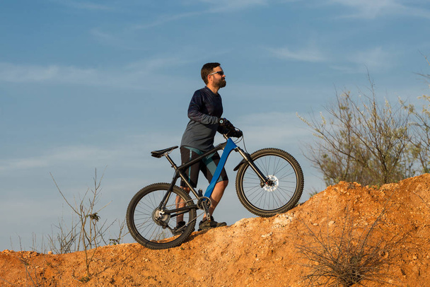 Ciclista en pantalones cortos y jersey en una moderna bicicleta rígida de carbono con una horquilla de suspensión neumática pasea fuera de la carretera en las colinas de color rojo anaranjado al atardecer en verano
 - Foto, Imagen