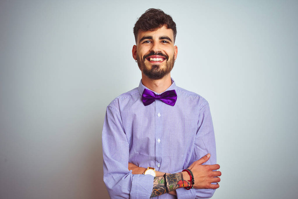 Joven hombre con tatuaje vistiendo camisa púrpura y pajarita sobre fondo blanco aislado cara feliz sonriendo con los brazos cruzados mirando a la cámara. Persona positiva
. - Foto, Imagen