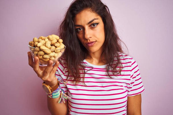 Nuori kaunis nainen tilalla kulho maapähkinöitä yli eristetty vaaleanpunainen tausta luottavainen ilme fiksu kasvot ajattelu vakava
 - Valokuva, kuva