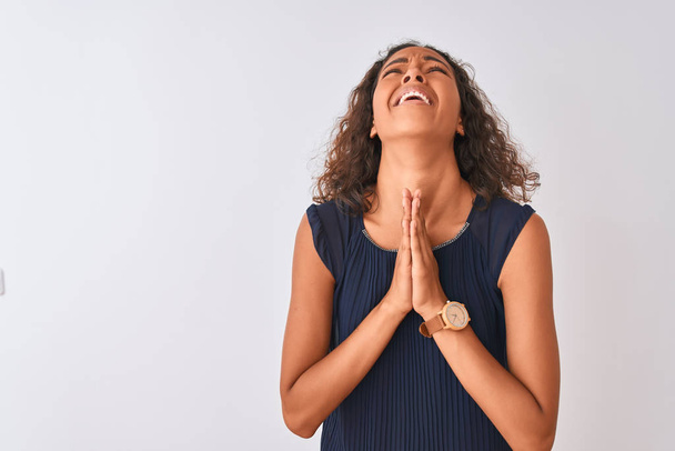 Fiatal brazil nő visel kék ruha állt át elszigetelt fehér háttér koldulás és imádkozik kézzel együtt remény kifejezése az arcon nagyon érzelmi és aggódik. Kér bocsánatot. Vallás-koncepció. - Fotó, kép