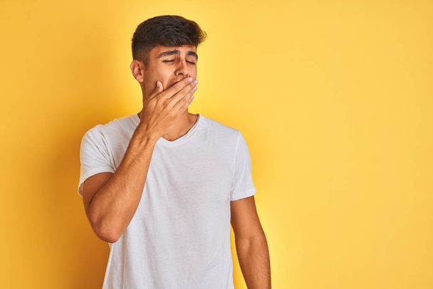 Homem indiano jovem vestindo camiseta branca em pé sobre fundo amarelo isolado entediado bocejo cansado cobrindo a boca com a mão. Inquieto e sonolência
. - Foto, Imagem