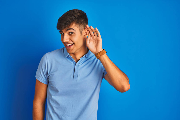 Jeune homme indien portant un polo décontracté debout sur fond bleu isolé souriant avec la main sur l'oreille en écoutant une rumeur ou des commérages. Concept de surdité
. - Photo, image