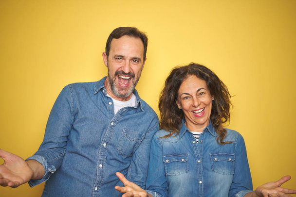 Beau couple d'âge moyen ensemble debout sur fond jaune isolé souriant gai avec les bras ouverts comme accueil amical, salutations positives et confiantes
 - Photo, image