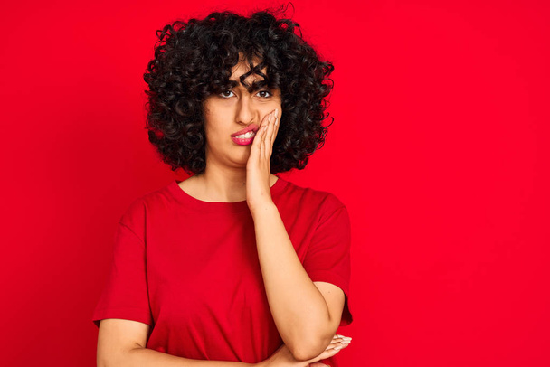 Mulher árabe jovem com cabelo encaracolado vestindo t-shirt casual sobre fundo vermelho isolado pensando que parece cansado e entediado com problemas de depressão com os braços cruzados
. - Foto, Imagem