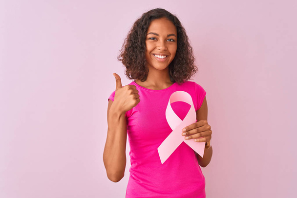 Young brazilian woman holding cancer ribbon standing over isolated pink background happy with big smile doing ok sign, thumb up with fingers, excellent sign - Foto, Bild