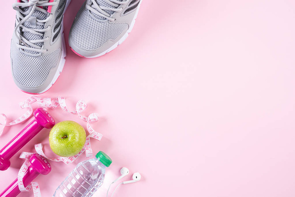 Healthy lifestyle, food and sport concept. Top view of athlete's equipment measuring tape pink dumbbell, sport water bottles, sport shoes and green apple on pink pastel background. - 写真・画像