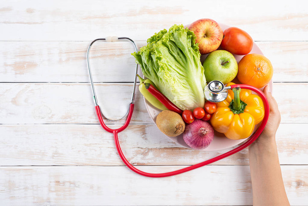Healthy lifestyle, food and nutrition concept. Close up doctor woman hand holding plate of fresh vegetables and fruits with stethoscope lying on white wooden table. - Photo, Image