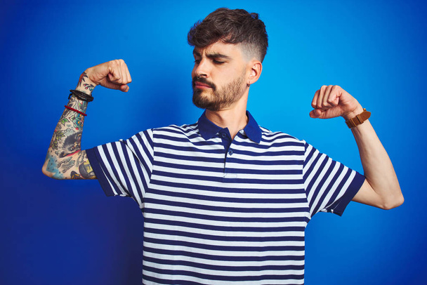 Joven hombre con tatuaje usando polo rayado de pie sobre fondo azul aislado mostrando los músculos de los brazos sonriendo orgulloso. Concepto Fitness
. - Foto, imagen