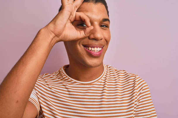 Jeune homme arabe beau portant t-shirt rayé debout sur fond rose isolé avec le visage heureux souriant faire ok signe avec la main sur l'œil regardant à travers les doigts
 - Photo, image