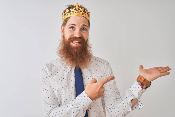 Jonge Redhead Ierse zakenman dragen kroon koning over geïsoleerde witte achtergrond verbaasd en glimlachend naar de camera tijdens het presenteren met de hand en wijzend met de vinger. - Foto, afbeelding