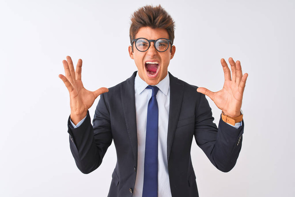 Young handsome businessman wearing suit and glasses over isolated white background crazy and mad shouting and yelling with aggressive expression and arms raised. Frustration concept. - Zdjęcie, obraz